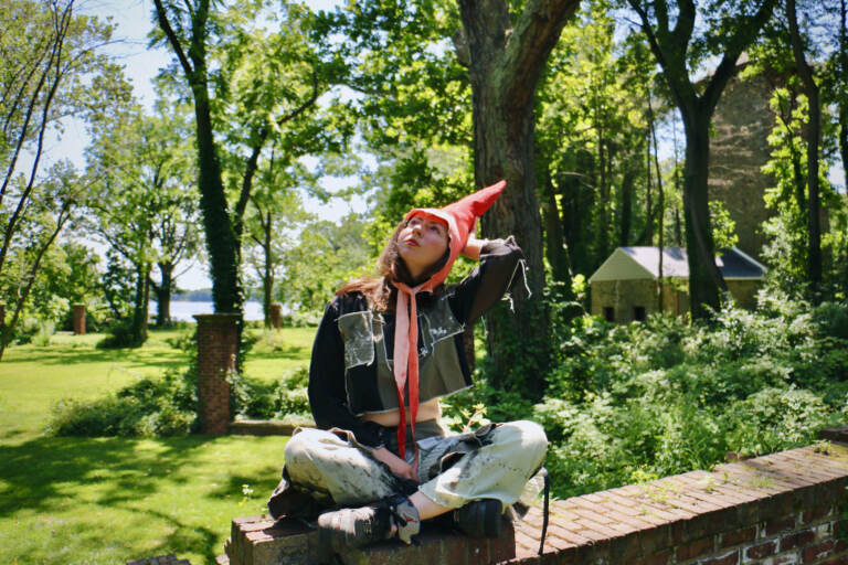 Artist in residence Alex Tatarsky, wearing a gnome costume, looks up towards the sky. Trees and grass are visible in the background.