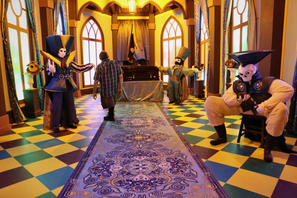 A room features strange court jester type figures and a tiled, elaborate floor, rug, and walls.