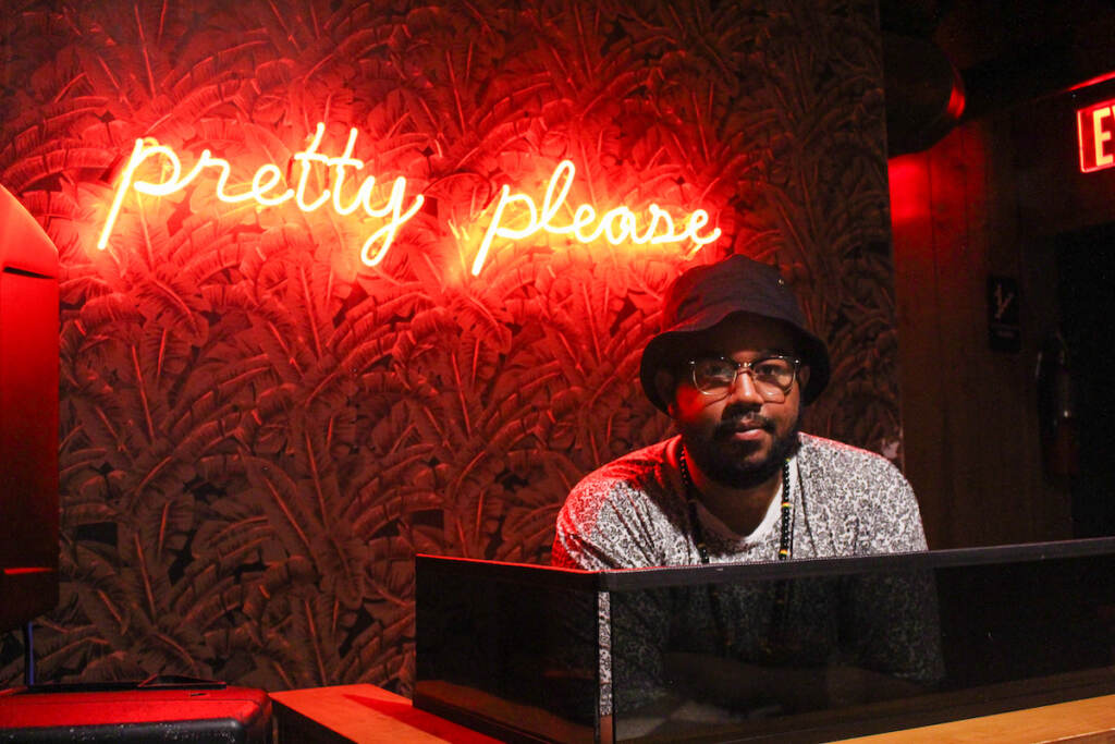DJ Rebel Foster poses for a photo. Behind him is visible a sign that reads "Pretty please"