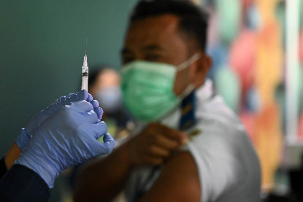 A booster vaccine is administered on the Indonesian resort island of Bali. A person holds up a vaccine syringe. A person in the background waits with their sleeve rolled up.