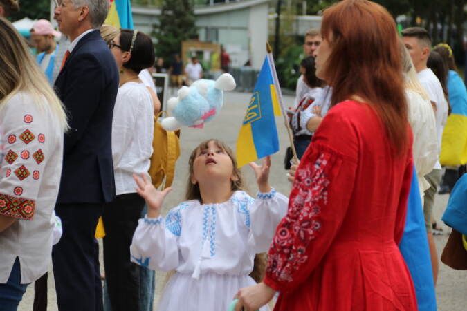 A child tossed a stuffed toy into the air. Next to her adults hold the Ukrainian flag.