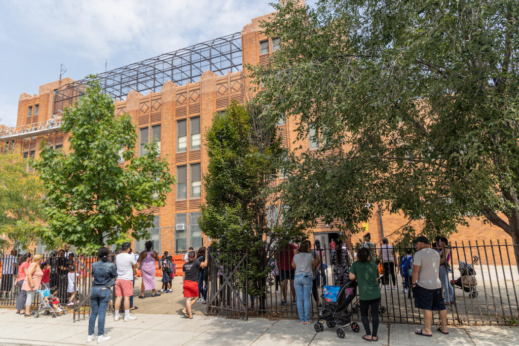 Parents wait in the shade for their children to be dismissed from school outside of the school building.