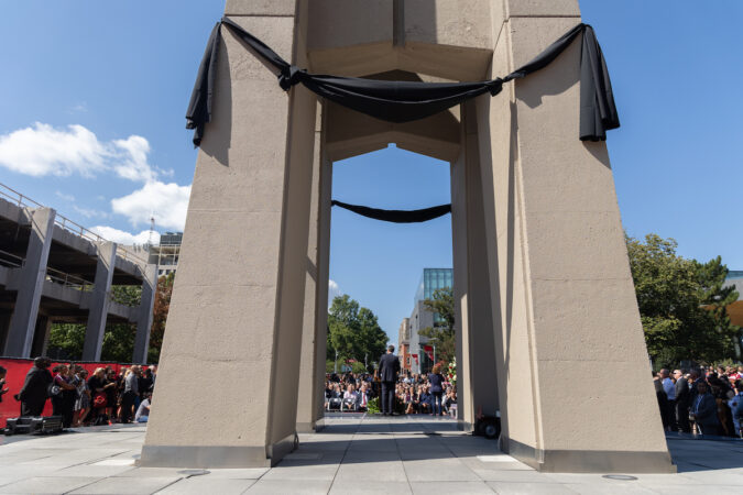 A black ribbon is draped over stone columns. In the distance Temple community members are gathered to mourn the loss of Joanne Epps.