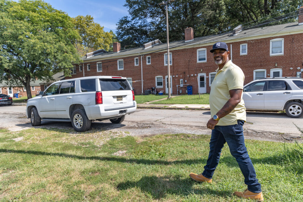 Leonard Williams looks forward, smiling, as he walks towards a street where two cars are parked. In the background are red-brick rowhomes.