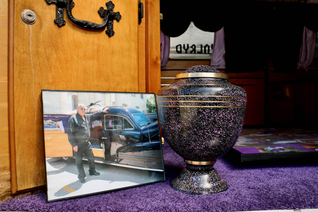 A photo of Russ Slegel next to an urn containing his ashes inside of the purple hearse he owned