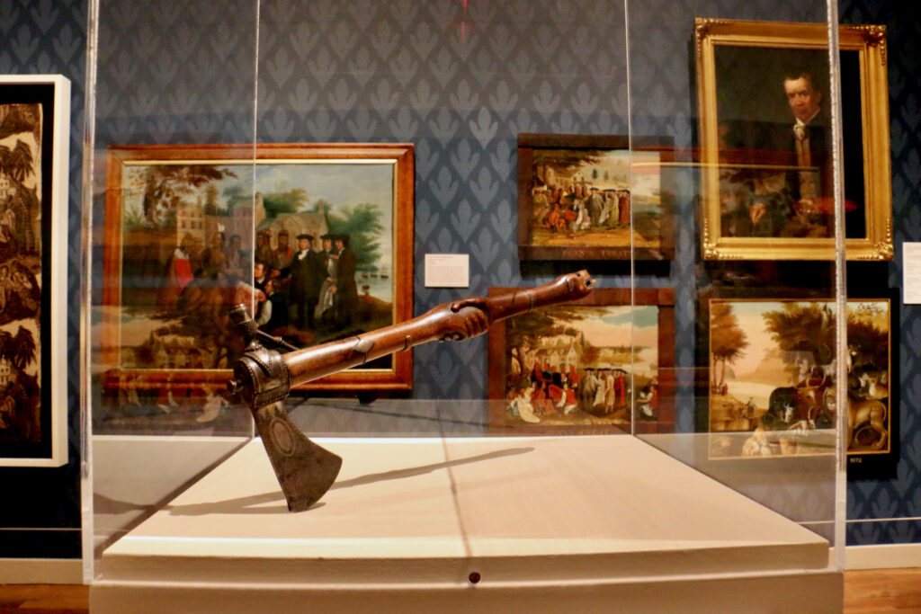 This pipe tomahawk made in the late 18th to early 19th century was likely intended to be presented during a treaty agreement. It is placed beside some of the many painted versions of the Penn Treaty with the Indians, an agreement that pushed the Lenape west
