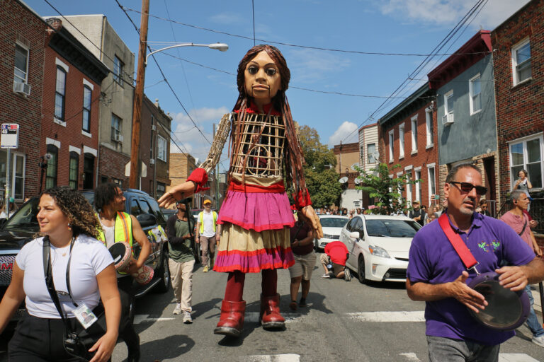 Little Amal, a 12-foot puppet, walks down a street in South Philadelphia, accompanied by residents and community members.