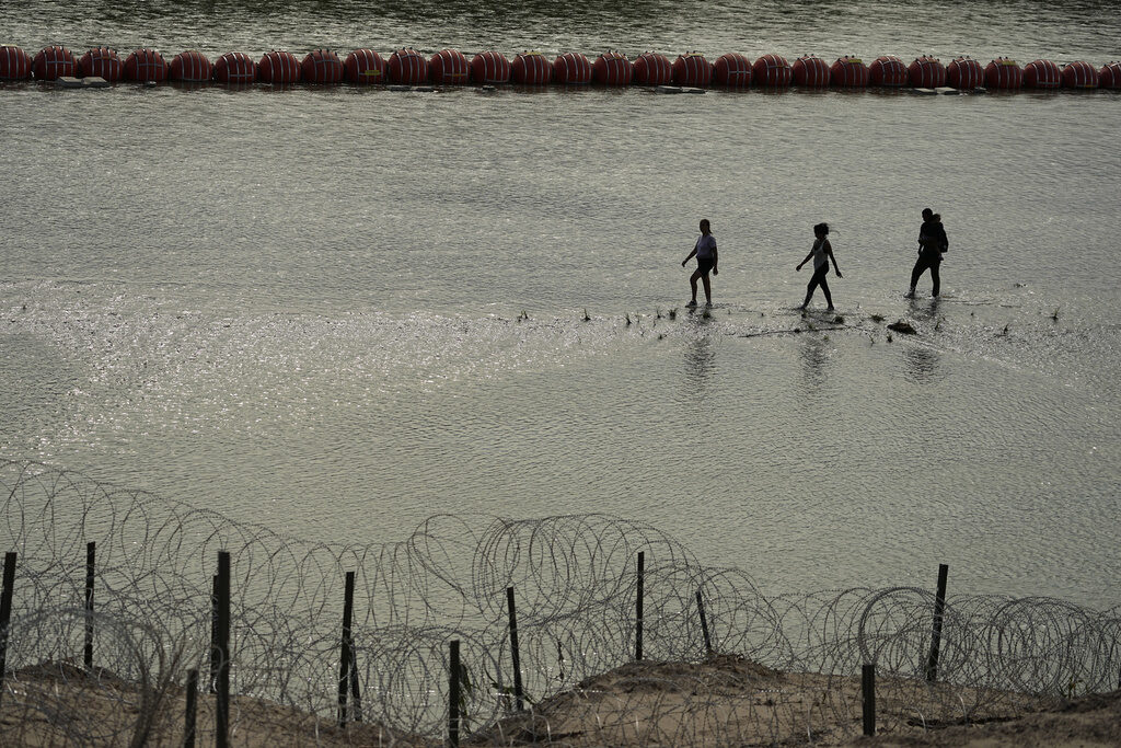 Migrants walk past large buoys being used as a floating border barrier on the Rio Grande, Monday, July 31, 2023, near Eagle Pass, Texas, as they cross from Mexico to the U.S. (AP Photo/Eric Gay)