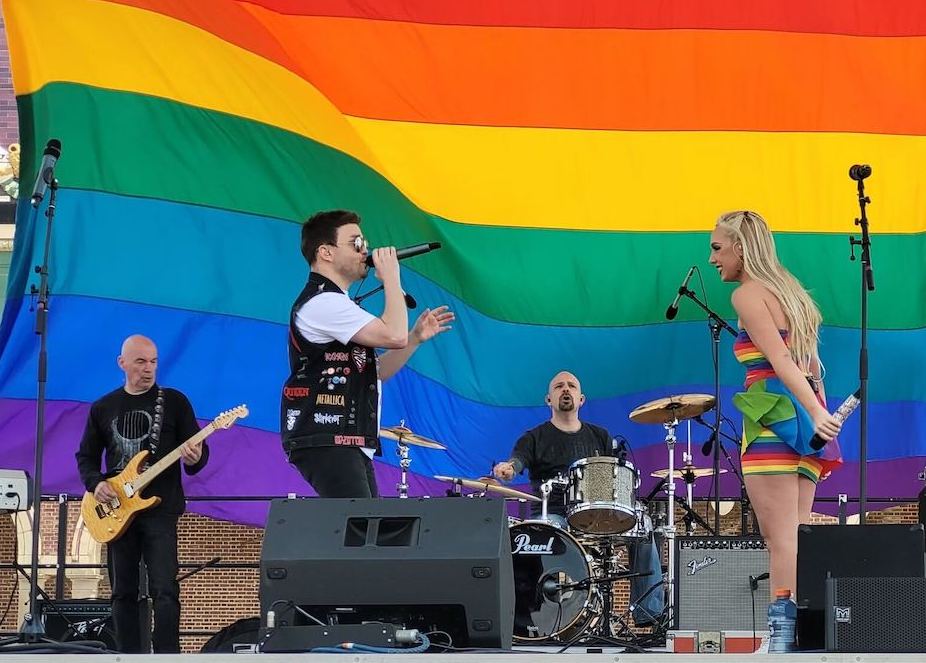 Kariselle-Snow-2 Kariselle Snow onstage as someone sings and a guitarist and drummer play in the background. In the back of the stage is a giant Pride flag.