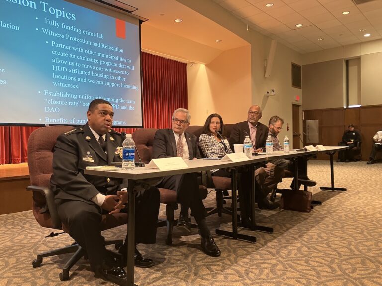 From left, Interim PPD Commissioner John Stanford, District Attorney of Philadelphia Larry Krasner and others participate in a panel, seated at a table, with a slideshow visible behind them.