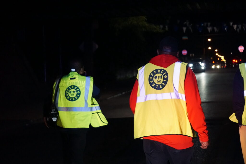 Two volunteers wearing bright yellow safety vests walk down a street.