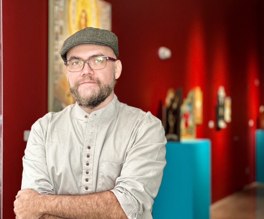 taller-puertorriqueno-exhibition-retablos-3 Patrick McGrath Muñiz poses for a photo. His paintings are visible behind him.