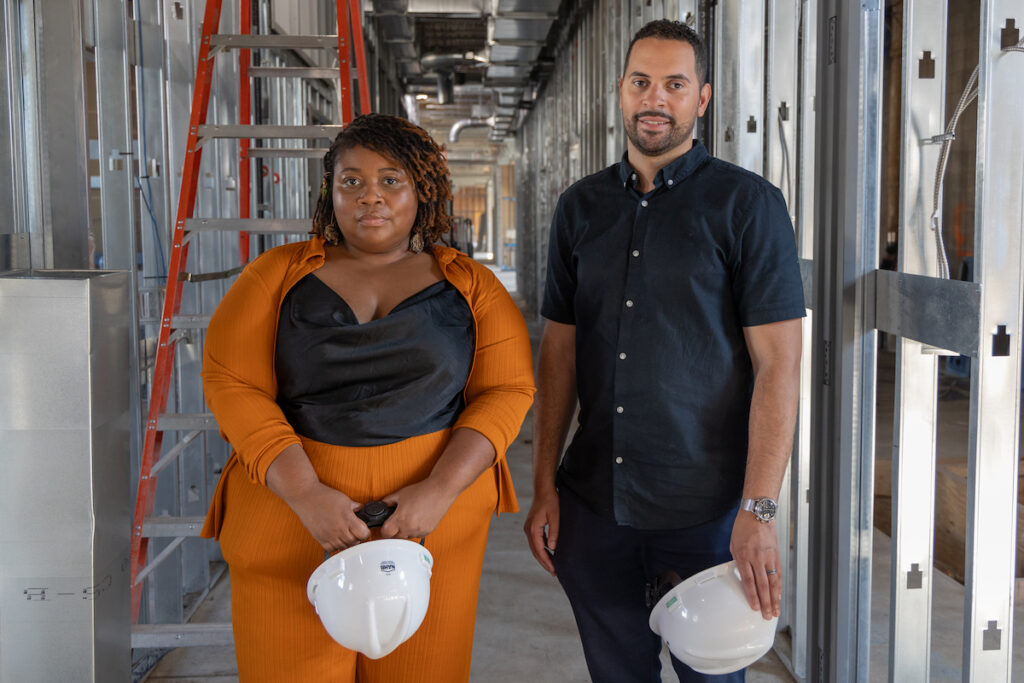 Tia Hall, left, and Alex Robles, right, pose for a photo in an area under construction, with hard hats in hand.