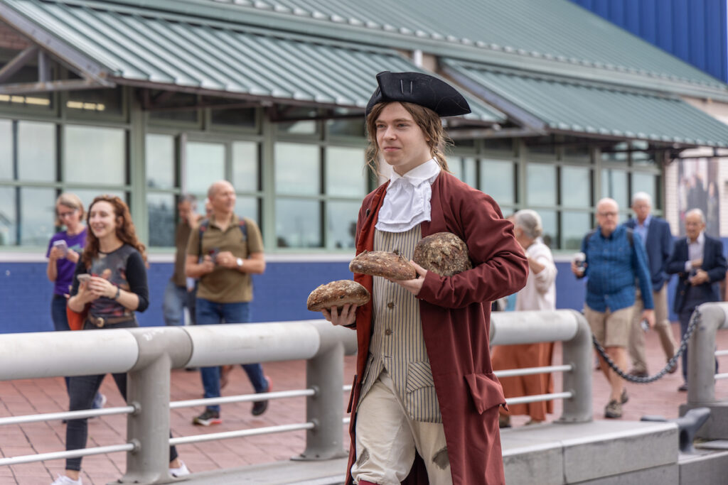 An actor playing Benjamin Franklin walks near the riverfront with three large bread loaves in his hands.