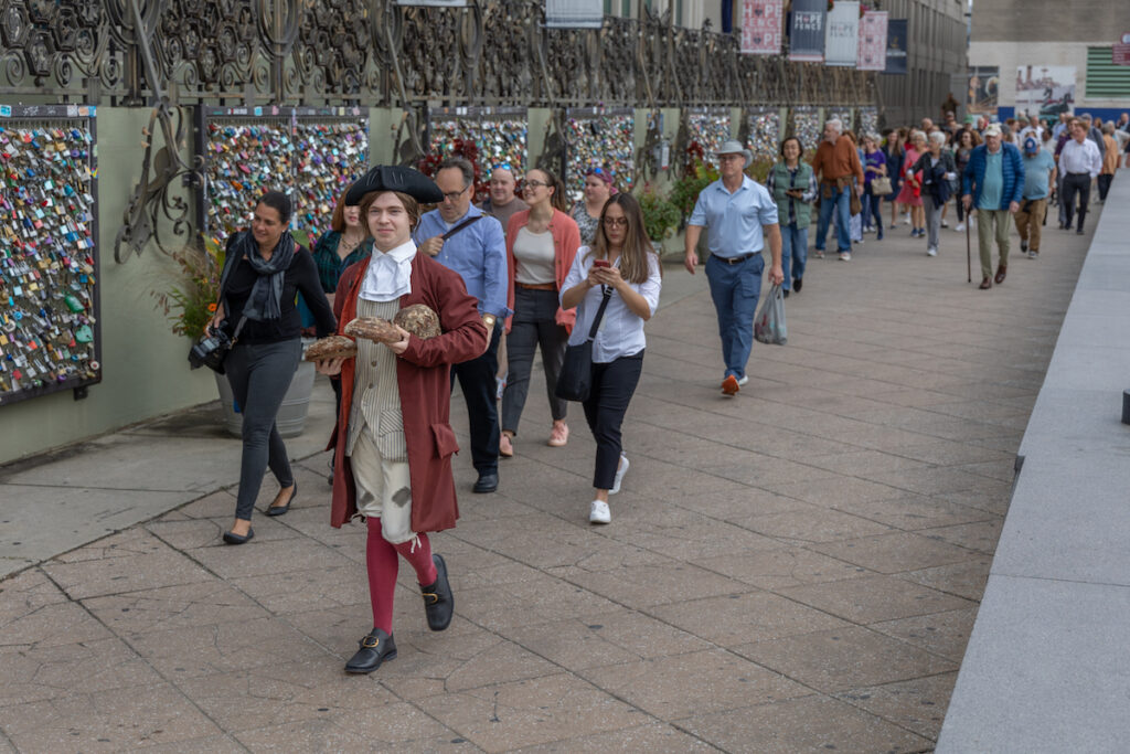 A Benjamin Franklin re-enactor walks down the street, followed by a crowd of spectators.