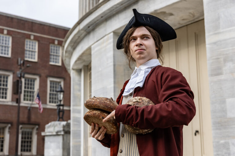 A Benjamin Franklin re-enactor, holding three bread loaves in his hands, looks into the distance