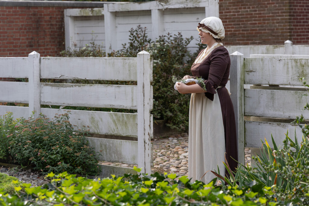 An actor playing Deborah Read, dressed in Revolutionary War-era clothing, stands in a garden.