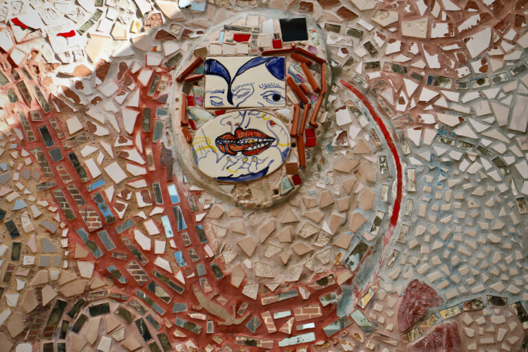 An up-close view of a detailed painted tile amid the larger mosaic on the side of a building wall.