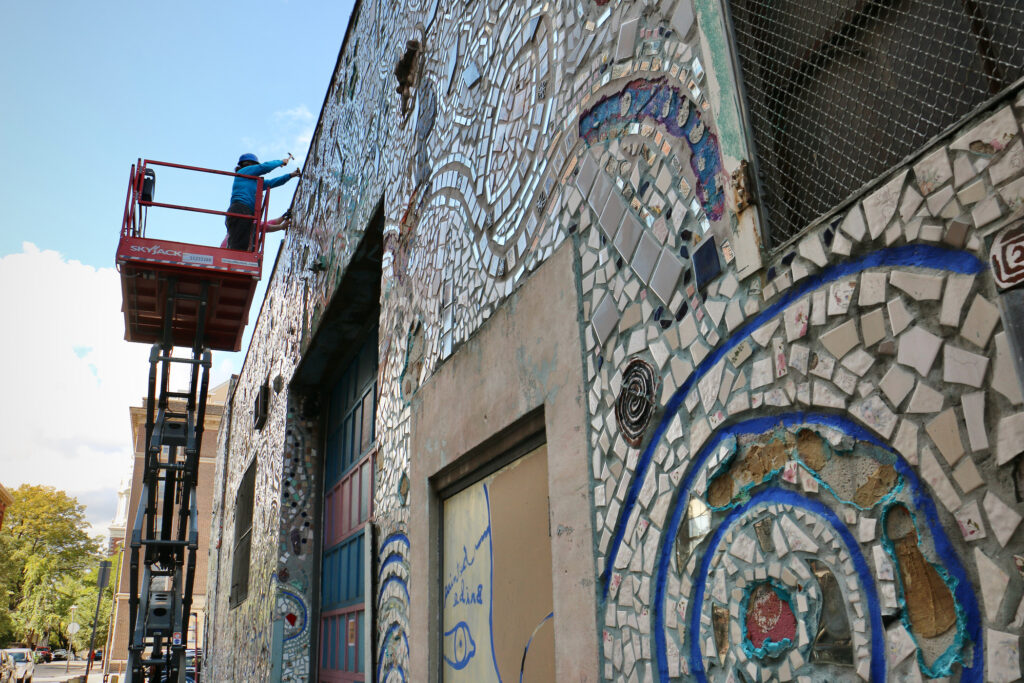 2023 10 17-e lee-stacey holder andrew jeffries-philadelphia old city painted bride art center-isaiah zagar mosaic Andrew Jeffries and Stacey Holder stand on a lift, chiseling pieces from a mural on a building.