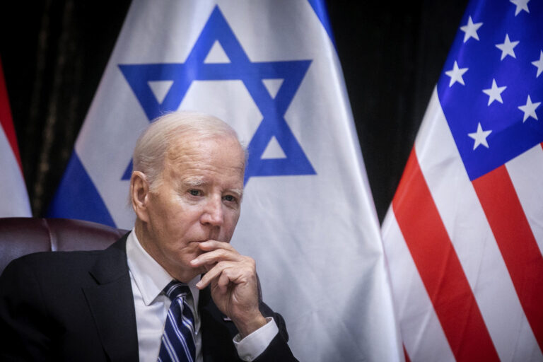 Biden sits in a chair, hand to face, with the Israeli and U.S. flags visible in the background.
