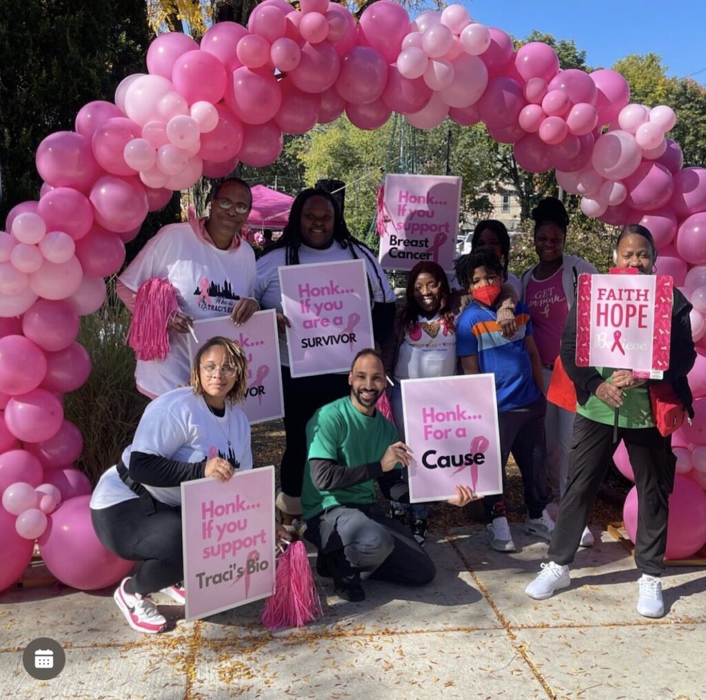 A group of people poses for a photo under an arbor of pink balloons holding signs reading Honk if you are a survivor.