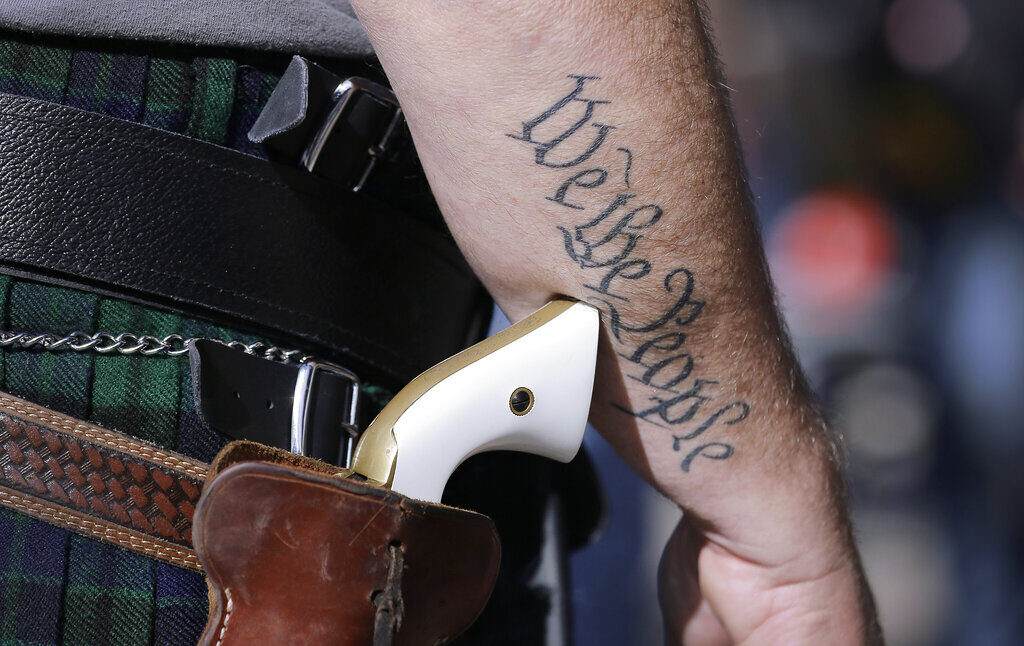 A close-up of someone's gun on a holster. On their arm is a tattoo that reads 