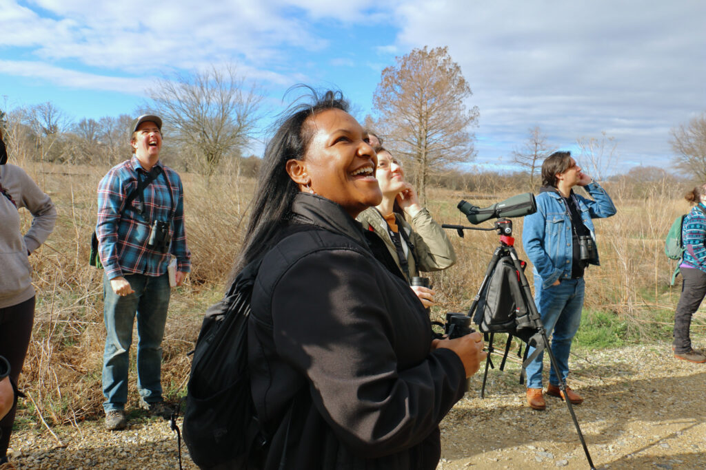 2022 03 20-e lee-nicole seahorne hameen-philadelphia fdr park-in color birding club (1) Nicole Seahorne Hameen looks upwards while holding a pair of binoculars. Other members of the bird watching group are visible in the background, also looking skyward.