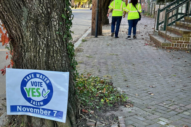 A sign reads Safe Water Vote Yes Salem November 7. In the background are two people walking down the sidewalk.