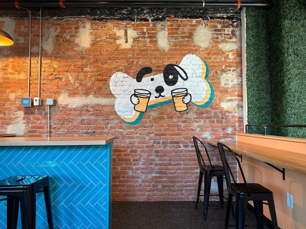boozymutt-01-scaled An image of a dog's face on a dog biscuit holding a couple of beers is painted on a brick wall behind a bar