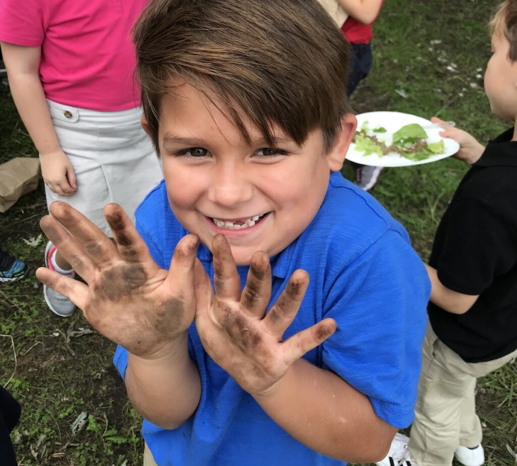 A student holds up his hands covered with dirt and smiles.
