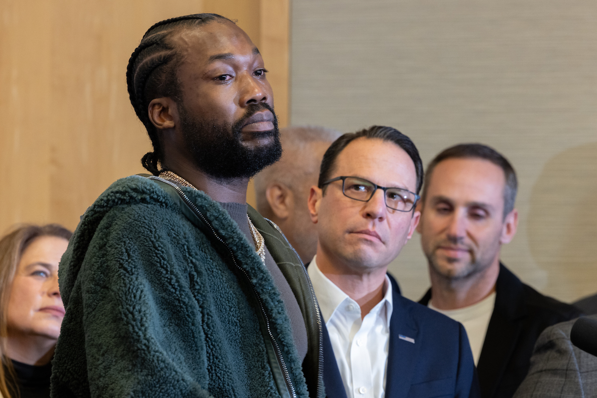 Meek Mill stands in front of a crowd of people, with Gov. Shapiro and other elected officials in the background