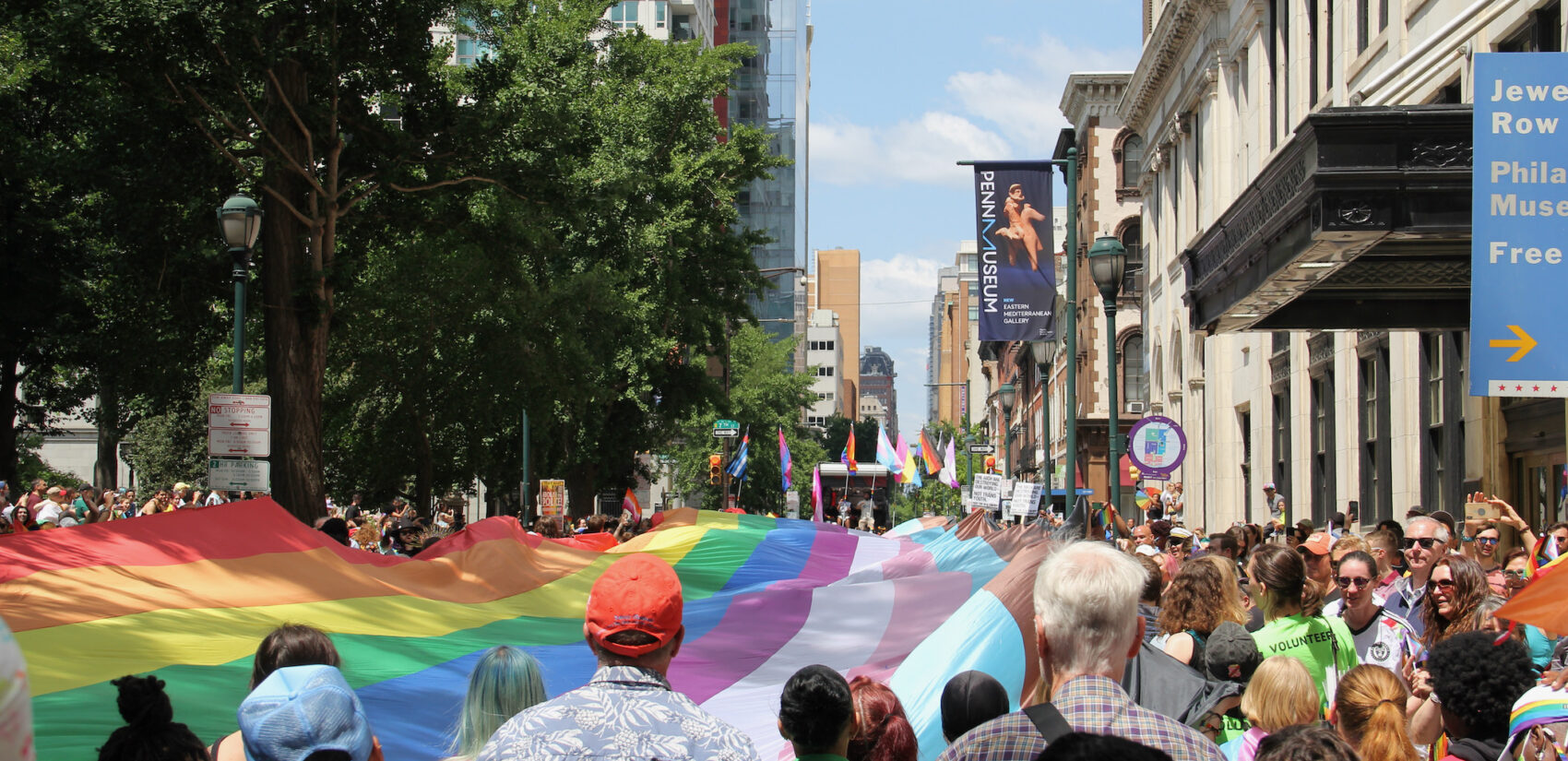 A large LGBTQ flag is carried through the streets by many people.