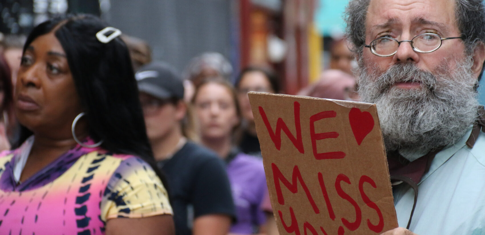 A man holds a handmade sign saying 'We Miss You'