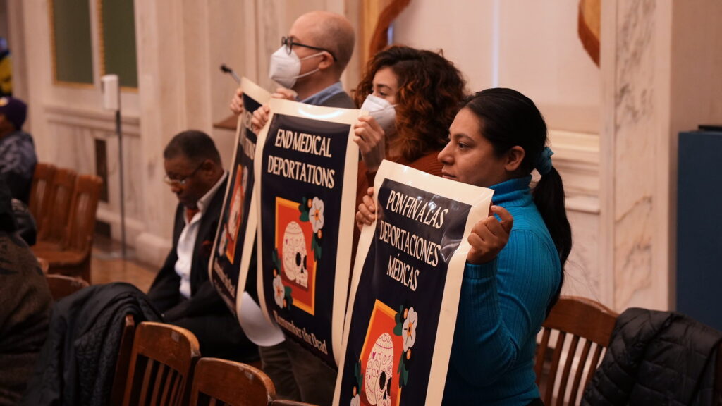 medical-deportations-2023-12-14-2 People holding up signs in City COuncil