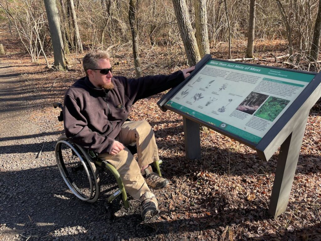Sean Holland, the Access Nature Disability Advocate for the Pinelands Preservation Alliance, points towards a sign on a trail.