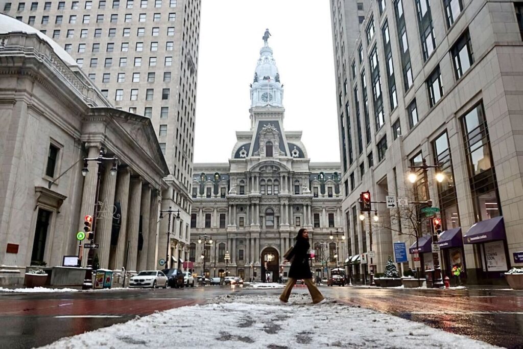 philly-snow-3 A person crossing the street in front of Philadelphia City Hall with snow on the ground.