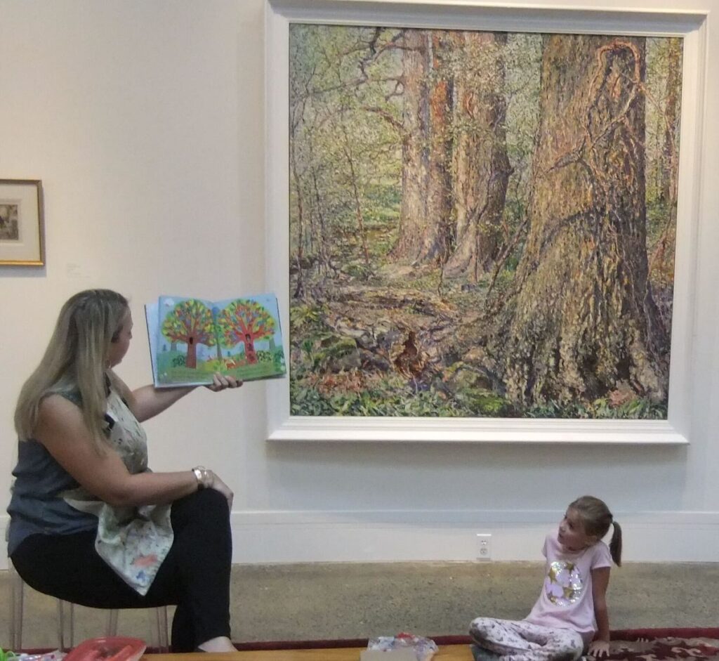 woodmere-art-museum-3 A person reads a picture book to children in front of a painting of a tree