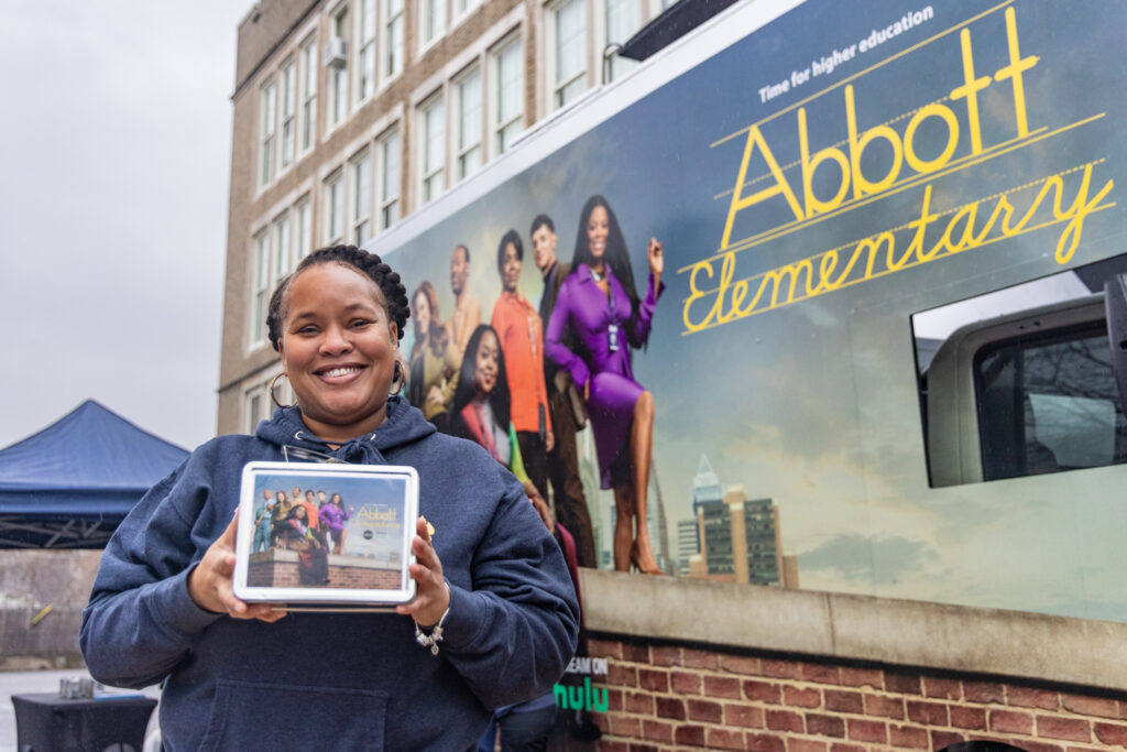 Ebonie Thorpe, a 6th grade teacher at the James R. Ludlow School in Philadelphia, holds up a lunchbox in front of an advertisement for Abbott Elementary