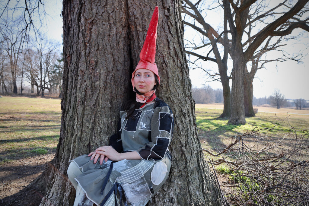 Alex Tatarsky, in gnome attire, sits at the foot of a tree