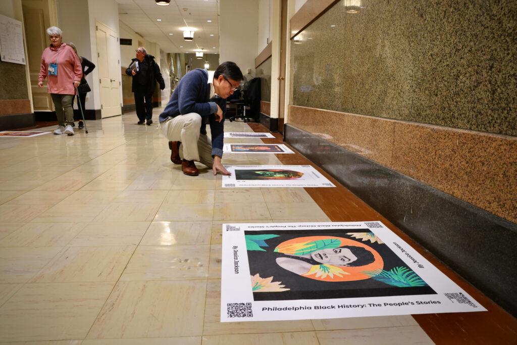 Tu Huynh, program manager for the Philadelphia Office of Arts, Culture, and the Creative Economy, kneels beside a portrait of Philadelphia artist and educator Moe Brooker