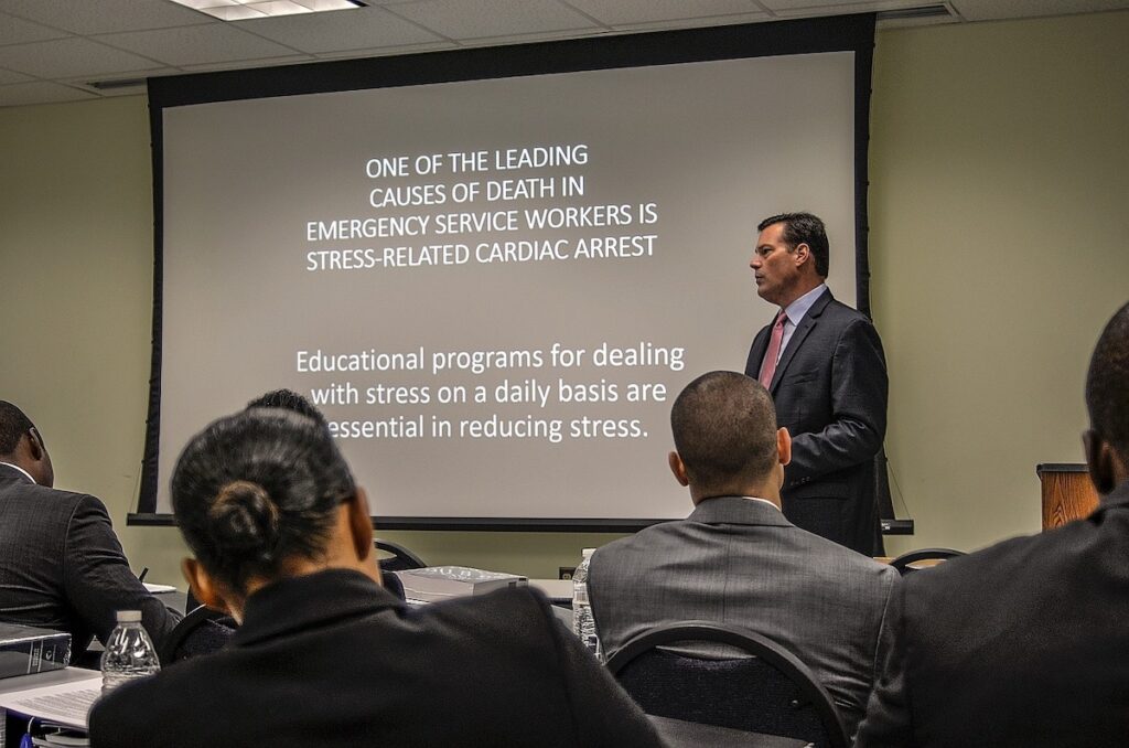 A person speaks in front of a group of people with a slideshow in the background that talks about stress-related cardiac arrest for emergency service workers.