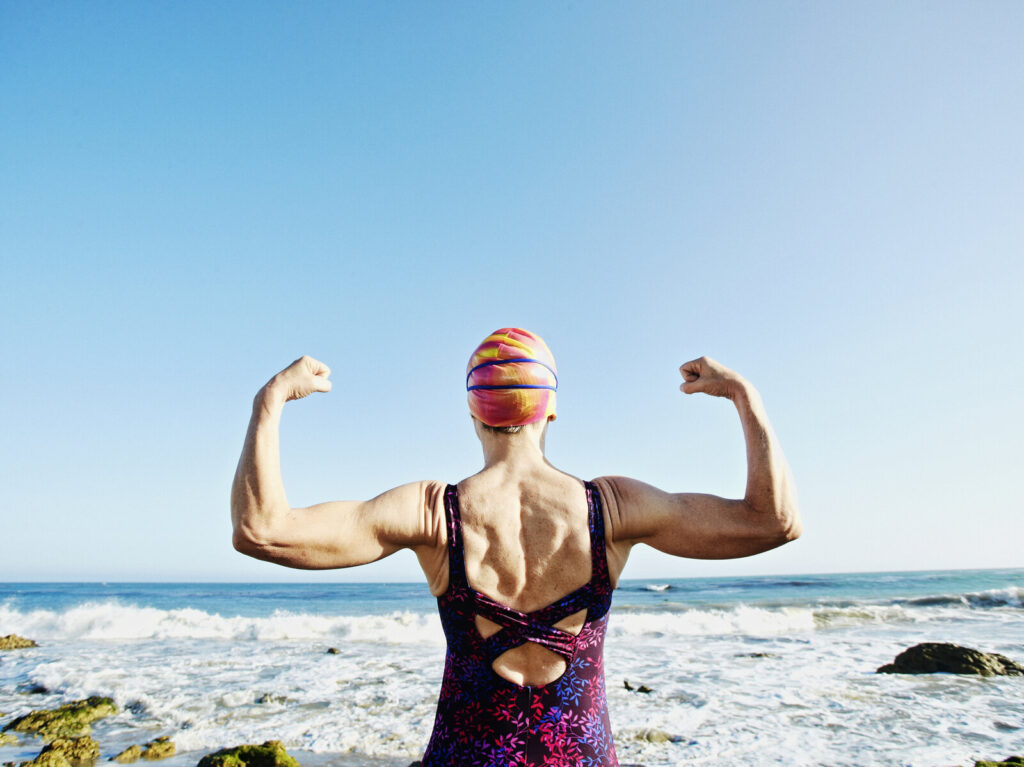 Older Caucasian woman flexing her muscles on beach A woman flexes her arms before entering the ocean
