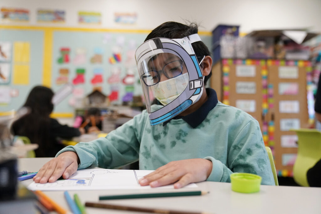COVID Isolation Changes Education A student wearing a mask and a face shield sits at a desk.