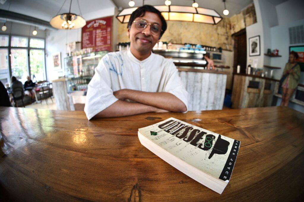 Nikil Saval posing for a photo in a coffeeshop with the book 'Ulysses'