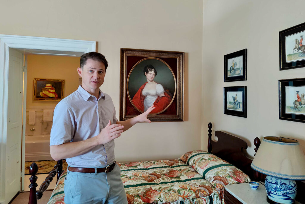 2024 07 19-p crimmins-john vick-andalusia pa historic house-portrait of adele sigoigne A man looks forward at the camera while gesturing toward a painting of a woman from the 19th century