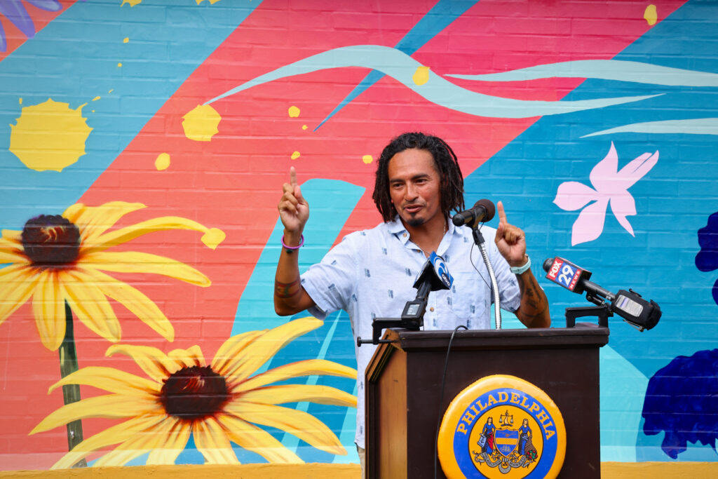 2024 07 31-e lee-calo rosa-philadelphia fitler square markward recreation center-mural artist A man stands at a podium in front of a mural on a wall featuring yellow flowers and varying colors