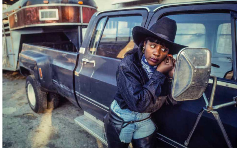A woman in western garb adjusts her earring in a car's side view mirror.