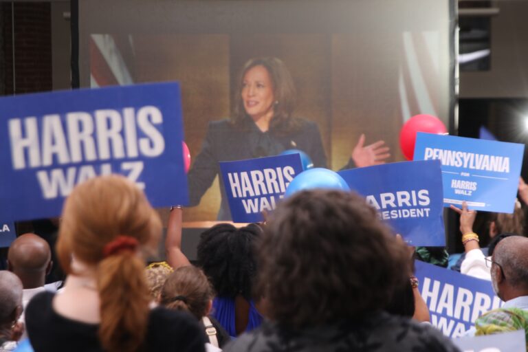 a projection screen shows Kamala Harris speaking, with viewers watching the screen