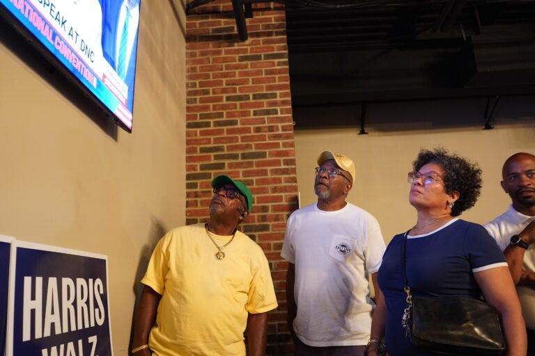 Attendees stand by and watch the TV