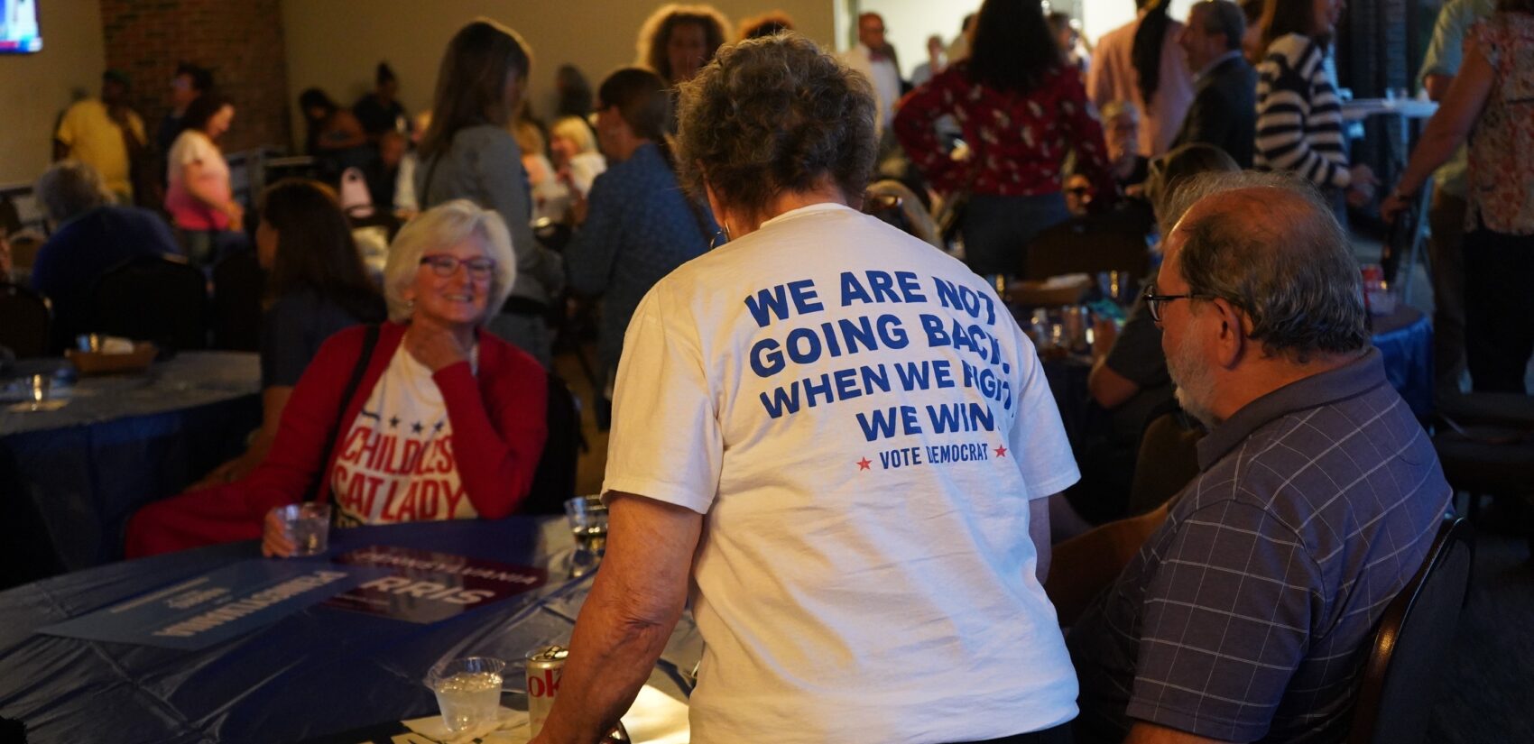 A person wearing a shirt reading WE ARE NOT GOING BACK stands by a table